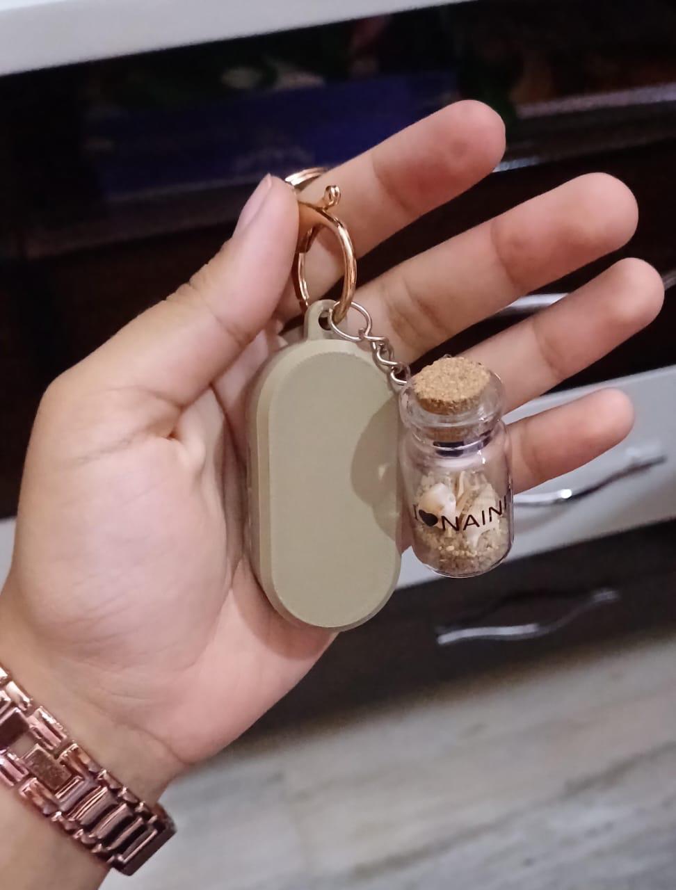 A hand holding a keychain carrying a smooth taupe capsule enclosure beside a small souvenir bottle