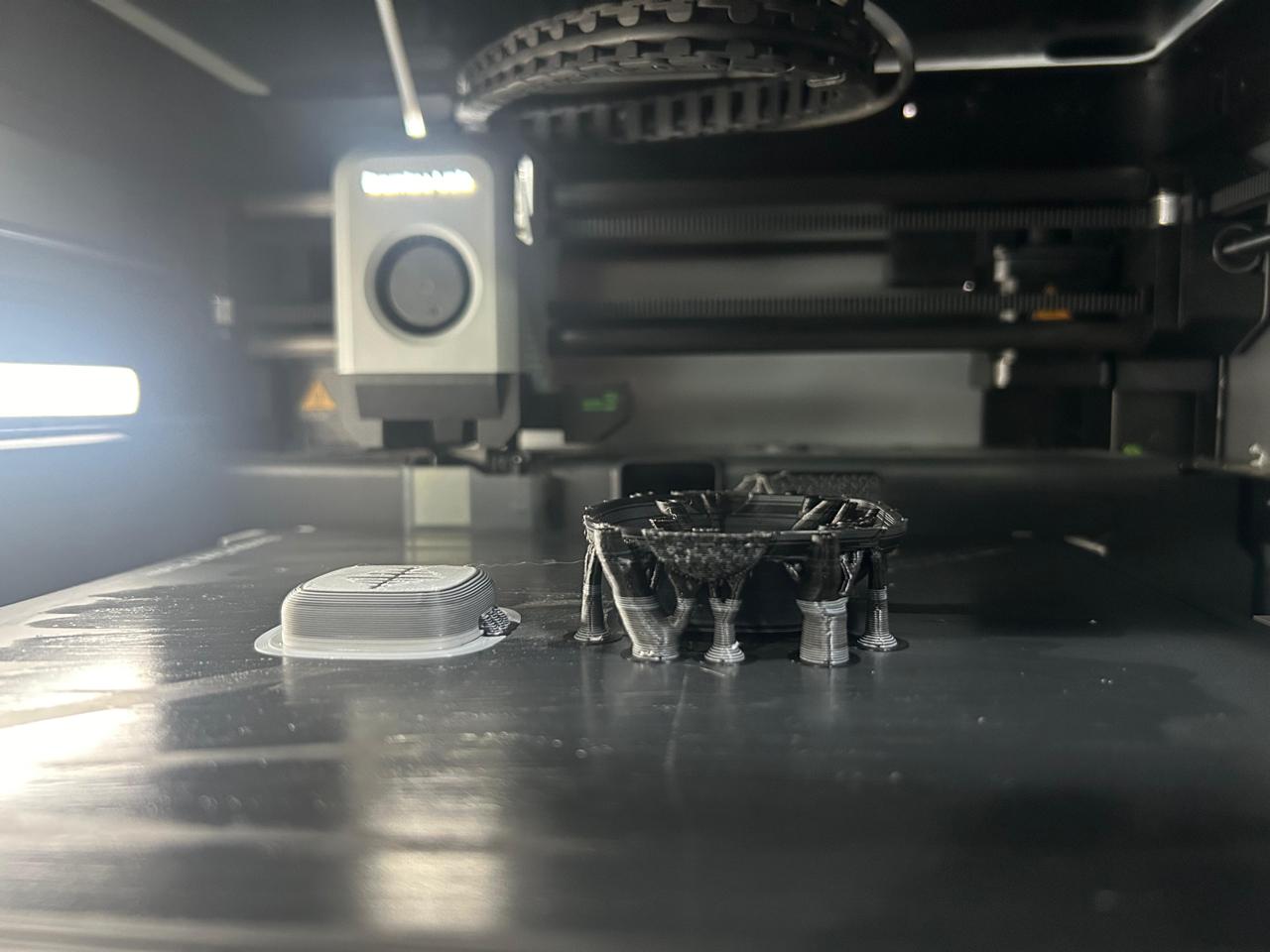Inside a 3D printer: a curved black printed shell held up by tree supports, beside a small light-grey printed piece