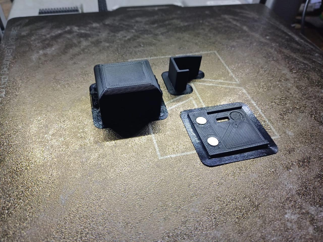 Three black 3D-printed parts on a printer build plate: a hollow enclosure piece, a small bracket and a flat plate holding two circular magnets