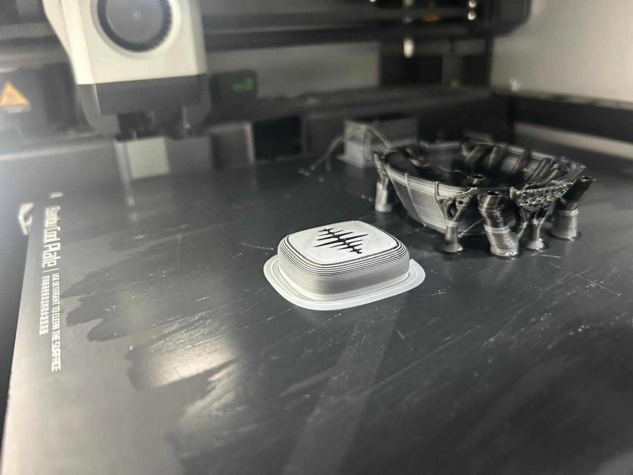 A 3D printer build plate from above: a light-grey printed piece with an embedded sound-wave graphic, and a curved black shell resting on tree supports