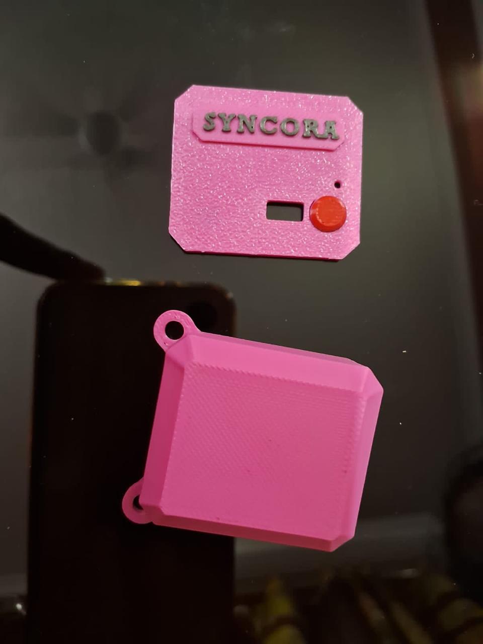 Two pink 3D-printed enclosure pieces on a dark surface: a lid embossed SYNCORA with a red push button, and a casing with two strap eyelets